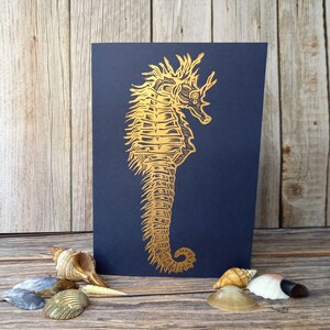 May include: A gold foil seahorse illustration on a dark blue card. The card is surrounded by seashells.