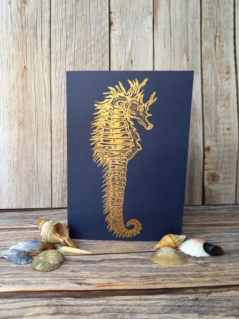 May include: A gold foil seahorse illustration on a dark blue card. The card is surrounded by seashells.