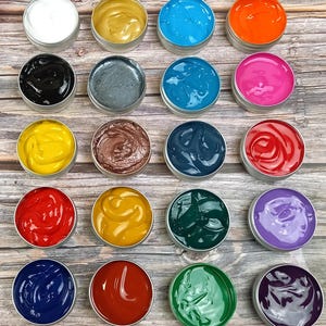 May include: A collection of small, round metal tins filled with colorful, creamy substances. The colors include white, gold, blue, orange, black, gray, teal, pink, yellow, brown, navy, red, and green. The tins are arranged on a wooden surface.