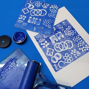 May include: A blue ink pad, a blue roller, and two white sheets of paper with blue ink patterns. The patterns are geometric shapes and dots. The text "FINE" is printed on one of the sheets.