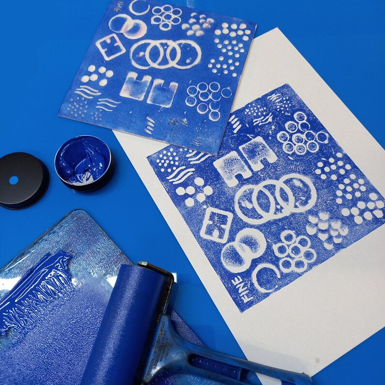 May include: A blue ink pad, a blue roller, and two white sheets of paper with blue ink patterns. The patterns are geometric shapes and dots. The text "FINE" is printed on one of the sheets.