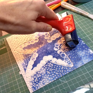 May include: A hand using a red handled roller to apply ink to a blue and white patterned linoleum block. The block features a star shape with a series of circles around the edges.