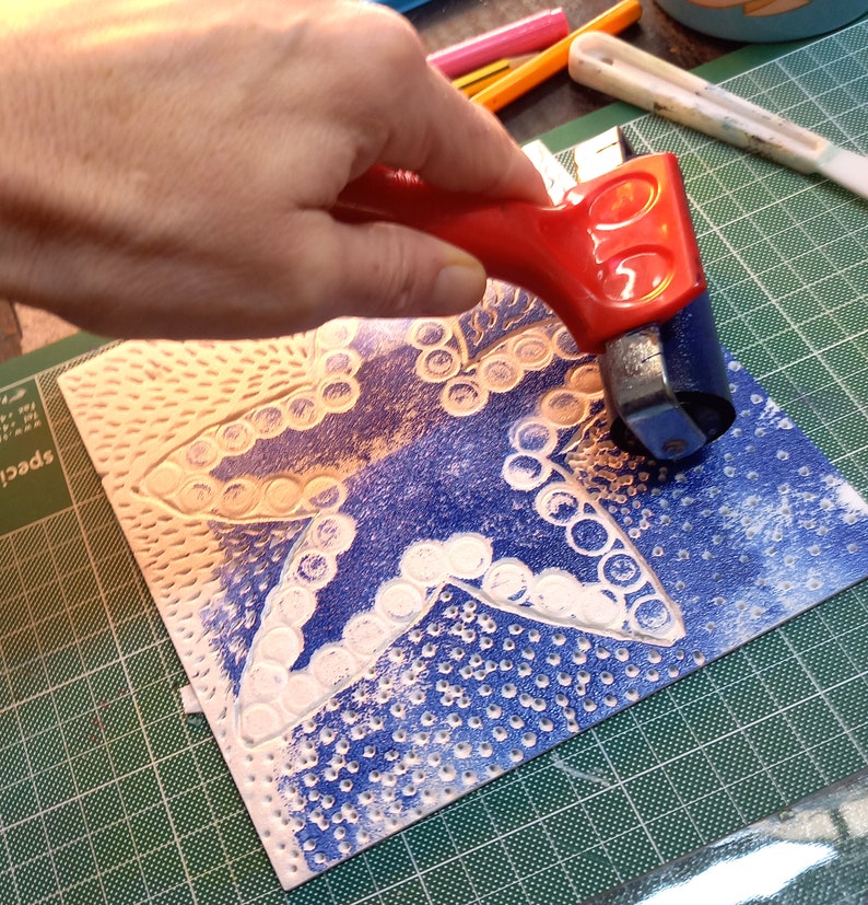 May include: A hand using a red handled roller to apply ink to a blue and white patterned linoleum block. The block features a star shape with a series of circles around the edges.