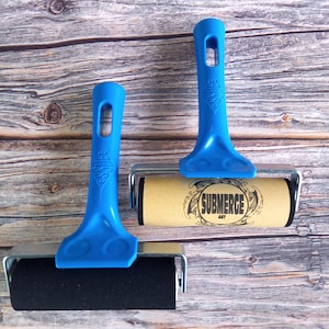 Op de afbeelding: Twee blauwe plastic drukrollen met metalen frames. Eén roller heeft een effen zwarte rubberen rol, terwijl de andere een beige rol heeft met de tekst SUBMERGE ART in een cirkelvormig ontwerp. Beide handvatten zijn voorzien van het Essdee-logo.