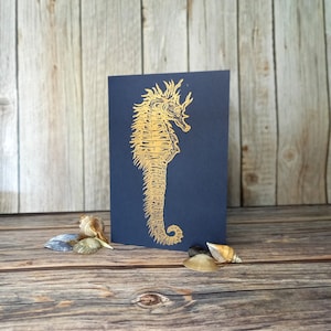 May include: A gold foil seahorse illustration on a dark blue card. The card is sitting on a wooden surface with seashells.