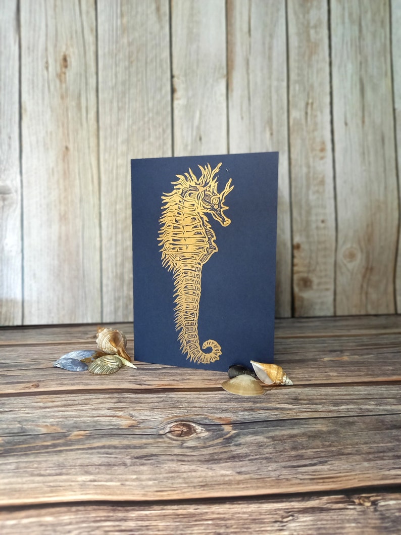 May include: A gold foil seahorse illustration on a dark blue card. The card is sitting on a wooden surface with seashells.