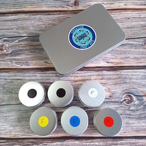 Op de afbeelding: Zes kleine ronde metalen containers met deksels. De deksels zijn gelabeld met zwarte, witte, gele, blauwe en rode cirkels. De containers zijn in een cirkel op een houten oppervlak gerangschikt. Een zilveren metalen doos met een blauw en wit logo dat "Creative Submerge Craft Kits" zegt, is op de achtergrond.