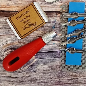 Peut inclure: Un couteau à linoléum à manche rouge avec une lame argentée et une boîte de lames de linoléum tranchantes. La boîte est marron avec les mots "CAUTION! sharp lino blades" et un dessin de main. Plusieurs lames argentées sont sur une surface bleue.