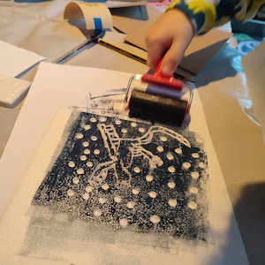 May include: A hand uses a red and black roller to apply white paint to a black and white print of a stylized creature with a dotted background.