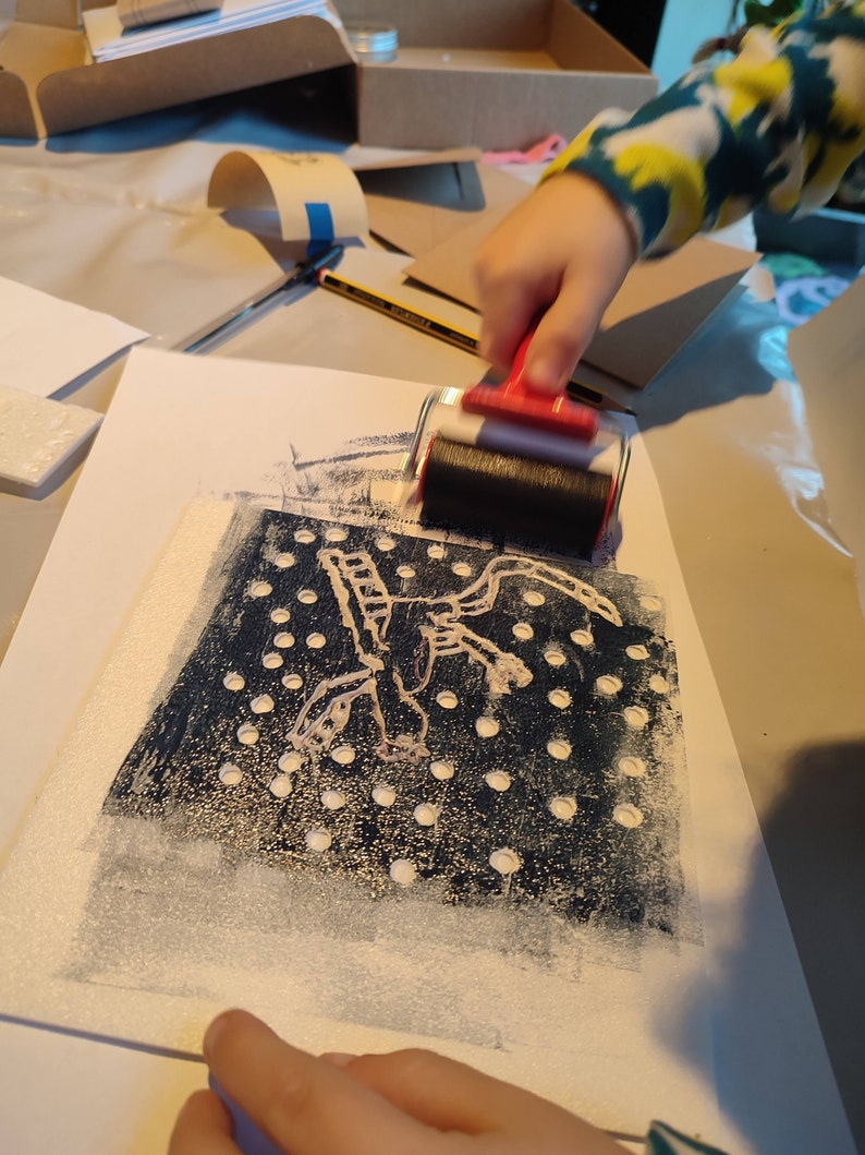 May include: A hand uses a red and black roller to apply white paint to a black and white print of a stylized creature with a dotted background.