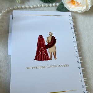 May include: A white spiral bound notebook with a gold line at the top and bottom. The cover features a watercolor illustration of a bride and groom in traditional Sikh wedding attire. The text "SIKH WEDDING GUIDE & PLANNER" is printed in black below the illustration.