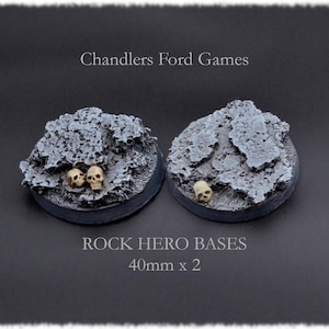 Könnte beinhalten: Zwei 40 mm runde Fels-Heldenbasen mit Schädeln. Die Basen sind grau bemalt mit einer rauen Textur.  "Chandlers Ford Games" und "ROCK HERO BASES 40mm x 2" sind auf dem schwarzen Hintergrund gedruckt.