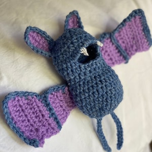 Può includere: Un giocattolo di pipistrello all'uncinetto fatto con filato blu e viola. Il pipistrello ha una piccola bocca e due gambe lunghe.