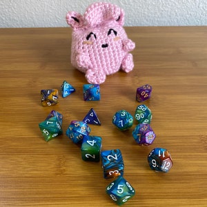 Può includere: Un peluche rosa all'uncinetto con un viso carino è seduto su una superficie di legno circondato da diversi dadi colorati con i numeri su di essi.