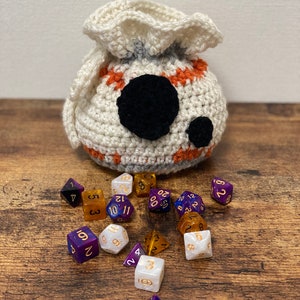 May include: A white crocheted dice bag with orange and gray accents and a black eye design. The bag is spilling out a variety of colorful dice with gold numbers.