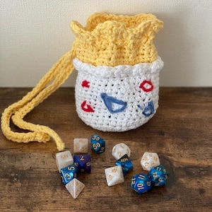 May include: A crocheted dice bag with a white face and yellow trim. The bag is open and spilling out a variety of colorful dice.