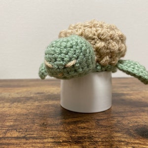 Può includere: Un giocattolo di tartaruga all'uncinetto con un guscio verde e una testa marrone. La tartaruga è seduta su un porta uova in ceramica bianca.