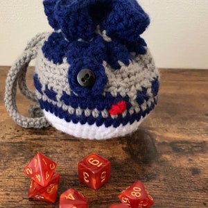 Puede incluir: Una bolsa de dados de crochet con un patrón azul y gris, un botón negro y una boca roja. Seis dados rojos con números dorados están esparcidos frente a la bolsa.
