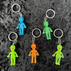 May include: A collection of alien-shaped keychains in various colors: blue, green, and orange. Each keychain features a small, translucent alien figure with large eyes and a silver keyring. The keychains are displayed on a dark, textured surface.