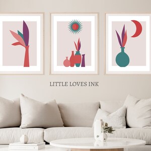 May include: Three framed abstract art prints with a pink background. The prints feature a stylized plant with a vase, a sun with a starburst, and a moon with a crescent shape.
