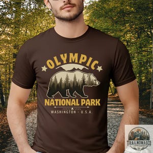 Puede incluir: Una camiseta marrón de manga corta con un gráfico de la silueta de un oso relleno con una escena de bosque. El texto OLYMPIC NATIONAL PARK WASHINGTON U.S.A. aparece en letras amarillas, junto a estrellas y formas de montañas sobre el oso.