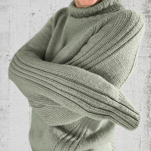 Könnte beinhalten: Ein salbeigrüner Strickpullover mit hohem Kragen und langen Ärmeln. Der Pullover hat eine gerippte Struktur.