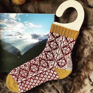 Könnte beinhalten: Eine rot-weiße gemusterte Socke mit einem senfgelben Bündchen, die an einem Holzbügel hängt. Die Socke ist mit einem geometrischen Muster gestrickt.