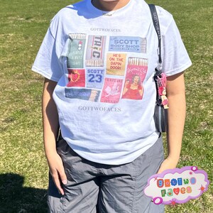 May include: White t-shirt with a graphic print of a collage of matchboxes. The matchboxes feature various images and text, including "Scott Body Shop", "He's on the Damn Door", "Scott 23", and "Karen's Cafe". The text "Got Two Faces" appears twice on the shirt.