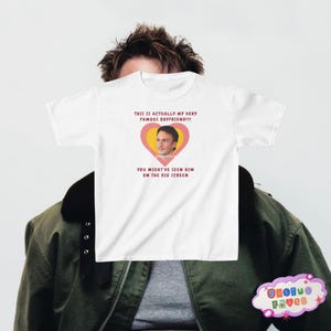 May include: White t-shirt with a pink heart graphic featuring a man's face and the text "THIS IS ACTUALLY MY VERY FAMOUS BOYFRIEND!!! YOU MIGHT'VE SEEN HIM ON THE BIG SCREEN".