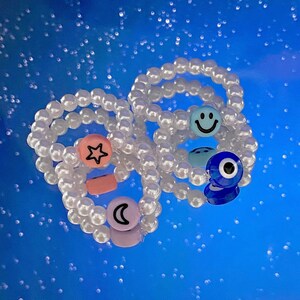 May include: Three white pearl beaded bracelets with colorful charms. The charms include a pink star, a pink crescent moon, a blue smiley face, and a blue evil eye.