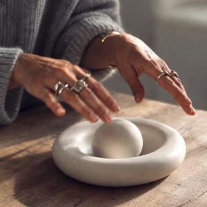 May include: A smooth, off-white circular decorative object featuring a thick, rounded rim and a central spherical element. The piece has a matte finish and a solid, uniform colour, presenting a clean, geometric form with a concave base area.