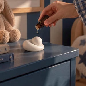 Könnte beinhalten: Ein kleiner, glatter, weißer Keramikdiffusor mit einer abgerundeten, kieselsteinartigen Basis und einer zentralen Kugel. Ein einzelner Tropfen klarer Flüssigkeit fällt aus einer kleinen, dunkel bernsteinfarbenen Glasflasche auf das Objekt.