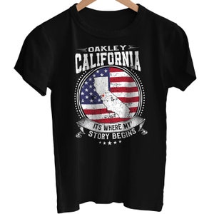 May include: Black t-shirt with a distressed white graphic of a California state outline superimposed on an American flag. The text "Oakley California" is above the graphic and "It's where my story begins" is below the graphic.