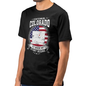 Puede incluir: Camiseta negra con un diseño gráfico blanco que presenta una bandera estadounidense desgastada con el estado de Colorado superpuesto. El texto "Centennial Colorado" y "It's Where My Story Begins" también está incluido en el diseño.