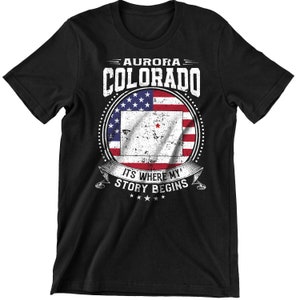 Może przedstawiać: Czarny T-shirt z białym nadrukiem graficznym przedstawiającym tekst "Aurora Colorado" i przetartą flagę amerykańską z czerwoną gwiazdą w środku. Tekst "It's where my story begins" znajduje się pod flagą.