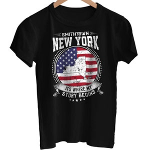 Puede incluir: Camiseta negra con un diseño gráfico blanco que presenta el texto "Smithtown New York" y un contorno del estado de Nueva York superpuesto a una bandera estadounidense. El gráfico también incluye el texto "It's where my story begins".