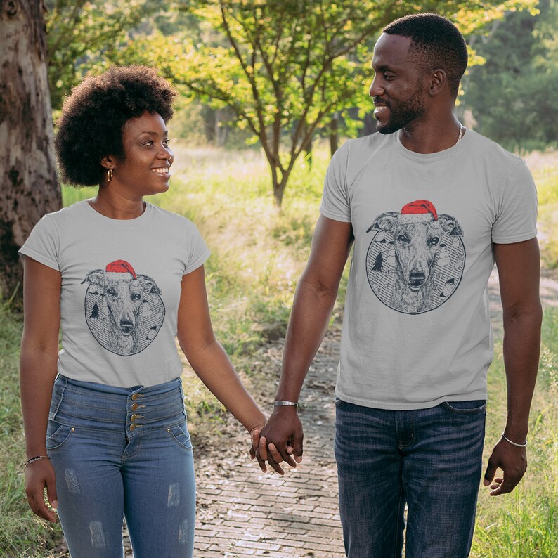 May include: A couple wearing grey t-shirts with a black and white illustration of a dog wearing a Santa hat. The dog is surrounded by a circle of pine trees. The couple is holding hands and walking in a park.