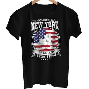 Op de afbeelding: Zwart T-shirt met een cirkelvormig ontwerp. Het ontwerp bevat de tekst "YONKERS NEW YORK" boven een afbeelding van de staat New York in de Amerikaanse vlag. De tekst "IT'S WHERE MY STORY BEGINS" staat eronder.