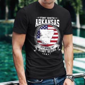 May include: Black t-shirt with a distressed graphic design featuring the state of Arkansas with a red, white, and blue American flag design. The text "Fort Smith Arkansas" and "It's where my story begins" is printed on the shirt.