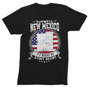Puede incluir: Camiseta negra con un gráfico de estilo vintage que presenta el texto "Roswell New Mexico" y una bandera estadounidense desgastada con el contorno del estado de Nuevo México. El gráfico también incluye el texto "It's Where My Story Begins".