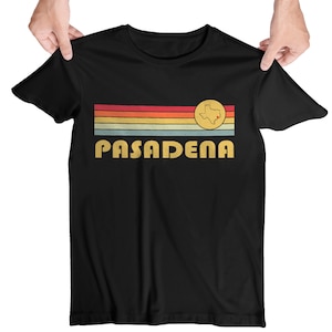 Może przedstawiać: Czarny T-shirt z retro wzorem w postaci tęczowych pasków i napisem "PASADENA" w żółtym kolorze. Mały okrąg z czerwonym punktem wewnątrz umieszczony jest na wzorze pasków, reprezentując stan Teksas.