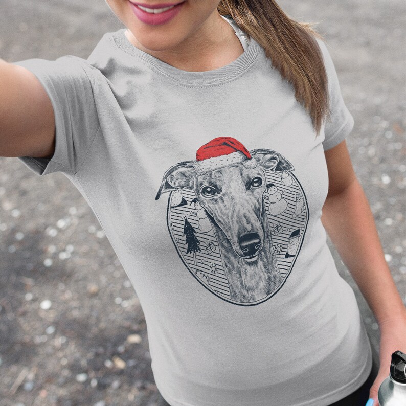 May include: A gray t-shirt with a graphic of a greyhound dog wearing a red Santa hat. The dog is surrounded by a circle of Christmas-themed illustrations, including a Christmas tree, ornaments, and snowmen.