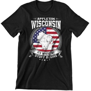 Może przedstawiać: Czarny T-shirt z białym nadrukiem graficznym przedstawiającym tekst "APPLETON WISCONSIN" i stylizowany obraz stanu Wisconsin nałożony na flagę amerykańską. Grafika zawiera również tekst "IT'S WHERE MY STORY BEGINS" z pięcioma gwiazdami poniżej.