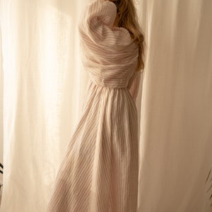 Pu&ograve; includere: Una donna indossa un lungo abito di lino bianco con sottili strisce rosa. L'abito ha la vita raccolta e maniche lunghe. &Egrave; in piedi davanti a una tenda bianca.