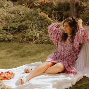 Pu&ograve; includere: Una donna con un vestito rosa a fiori &egrave; seduta su una coperta bianca in un'area erbosa. Si sta sistemando i capelli e ha un cestino da picnic e cibo nelle vicinanze.