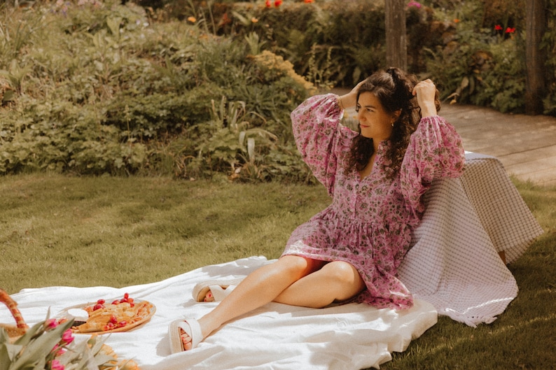 Pu&ograve; includere: Una donna con un vestito rosa a fiori &egrave; seduta su una coperta bianca in un'area erbosa. Si sta sistemando i capelli e ha un cestino da picnic e cibo nelle vicinanze.