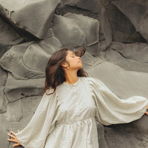 Pu&ograve; includere: Una donna con lunghi capelli scuri indossa una blusa a quadri bianchi e neri con maniche a sbuffo. &Egrave; seduta su una grande roccia grigia con gli occhi chiusi e la testa reclinata all'indietro.