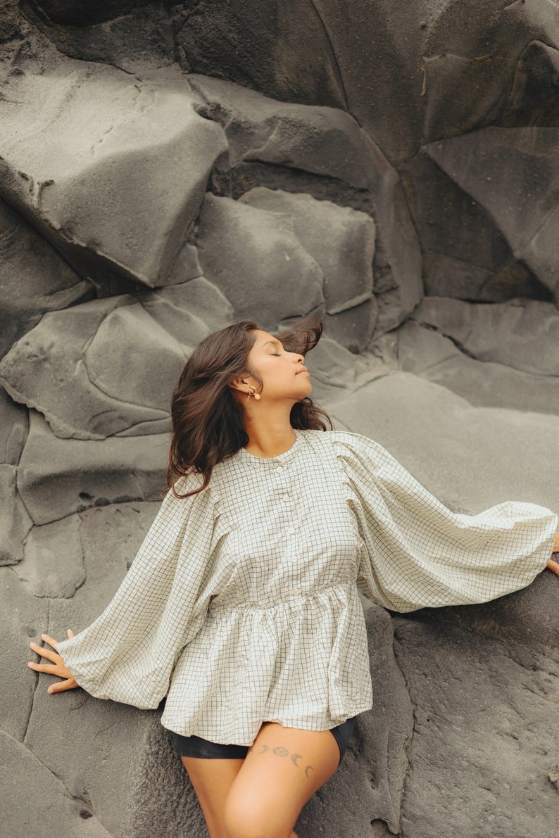 Pu&ograve; includere: Una donna con lunghi capelli scuri indossa una blusa a quadri bianchi e neri con maniche a sbuffo. &Egrave; seduta su una grande roccia grigia con gli occhi chiusi e la testa reclinata all'indietro.