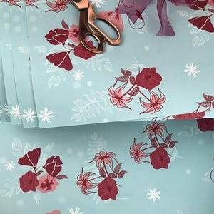 May include: A stack of light blue wrapping paper with a white floral pattern and red flowers. The paper is decorated with white snowflakes.