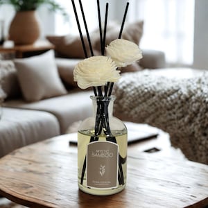 May include: A clear glass diffuser bottle with black reeds and two white flower-shaped decorations. The bottle contains a light yellow liquid and a label that reads "Mystic Bamboo". The diffuser sits on a round wooden table.